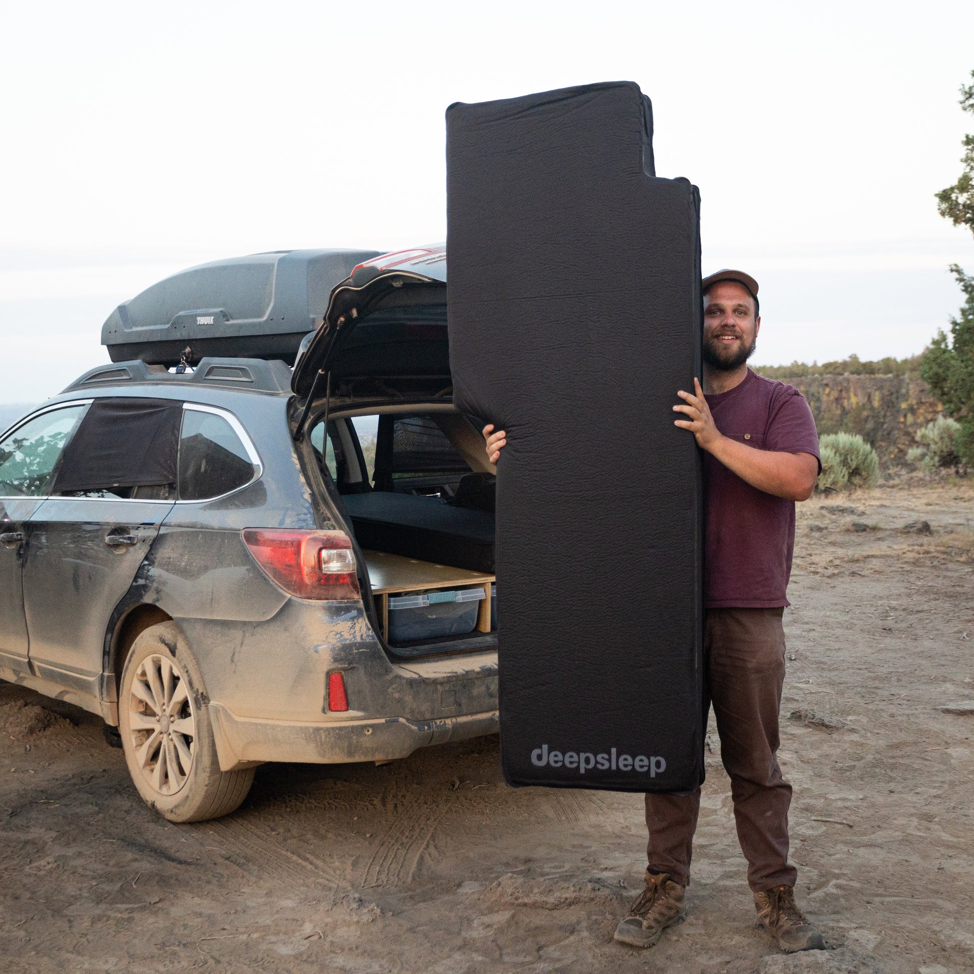 A man standing in front of a Subaru Outback SUV with a CarToCamp Deepsleep Solo Mat and a large piece of luggage.